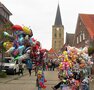 Das Bild zeigt Menschen auf der Kirmes vor dem Kirchturm Emsbüren und einen Luftballon- und Teddy-Stand